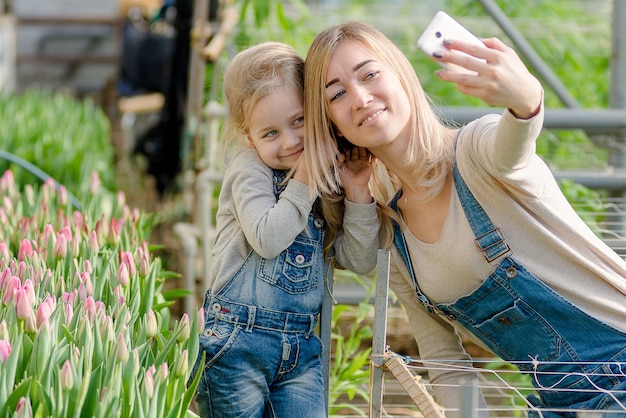 Una mujer con una hija pequeña se toma un selfie en un invernadero con flores en primavera.