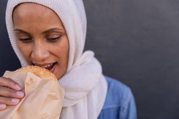 Mujer en hijab comiendo hamburguesa contra la pared