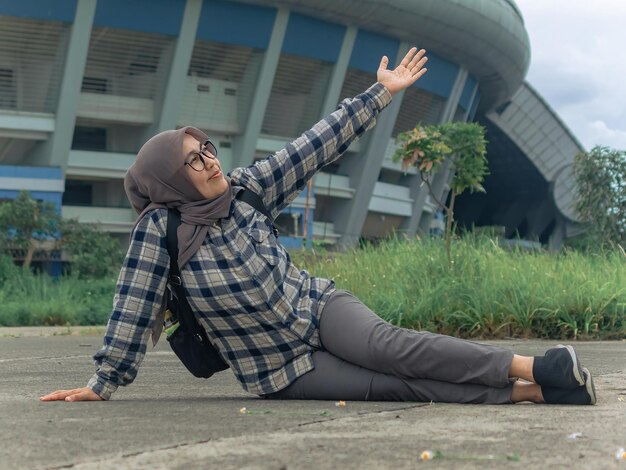 mujer en hijab con gafas sentada en el suelo con las piernas extendidas y mirando al cielo