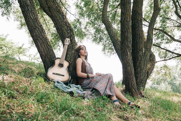 Mujer hippie con guitarra sentada cerca de un árbol en el bosque