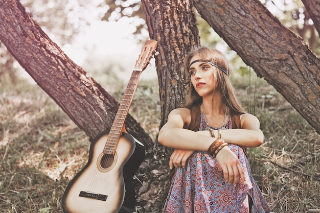 Mujer hippie joven pensativa sentada cerca de un árbol en el bosque