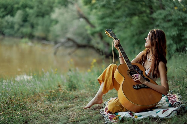 Una mujer hippie tocando su guitarra sonríe y canta canciones en la naturaleza sentada en una alfombra por la noche en la luz del sol poniéndose un estilo de vida en armonía con el cuerpo y la naturaleza foto de alta calidad