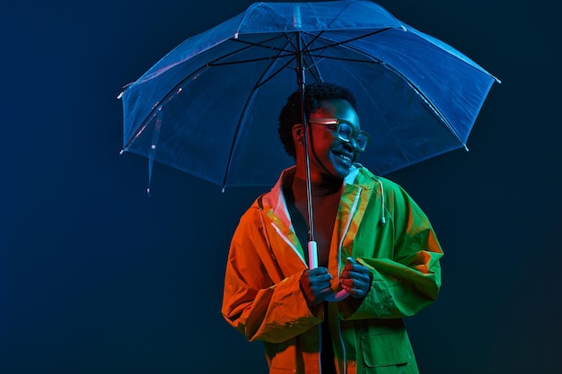 Mujer hipster en impermeable con paraguas transparente de pie en luz de neón sobre fondo de estudio oscuro