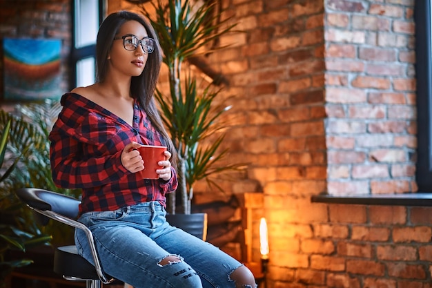 Una mujer hipster morena vestida con una camisa de lana y jeans bebe café en una habitación con interior de loft.