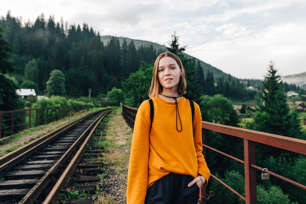 mujer hipster positiva con ropa informal caminando por la vía de un puente ferroviario de montaña