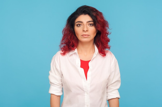 Mujer hipster seria y concentrada con cabello rojo elegante en camisa blanca mirando a la cámara con expresión determinada y confiada individualidad carismática tiro de estudio interior aislado en fondo azul