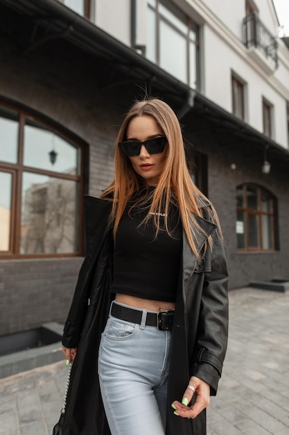 Mujer hipster sexy de moda con gafas de sol frescas en traje urbano negro de moda con camiseta de abrigo negro y jeans caminando en la ciudad