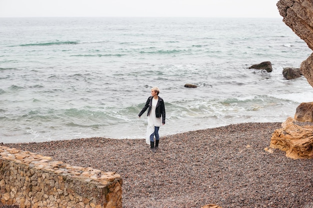 Mujer hipster con vestido y chaqueta caminando cerca del mar en otoño