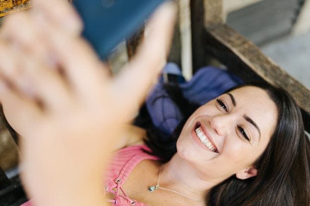 Foto mujer hispana alegre que usa el teléfono móvil mientras está acostado en un banco en el parque público - niña divirtiéndose mientras charla en el teléfono inteligente al aire libre - estilo de vida juvenil, redes sociales y concepto de tecnología