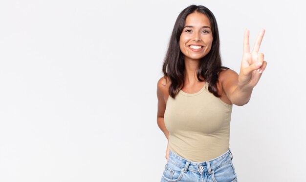 Mujer hispana bastante delgada sonriendo y mirando feliz, gesticulando victoria o paz