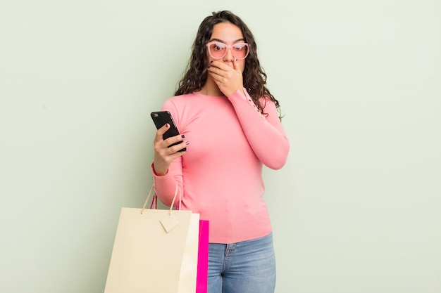 Mujer hispana bonita joven que cubre la boca con las manos con un concepto de bolsas de compras sorprendidas