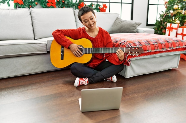 Mujer hispana con cabello corto aprendiendo guitarra clásica de un tutorial en línea que se ve positiva y feliz de pie y sonriendo con una sonrisa segura mostrando los dientes
