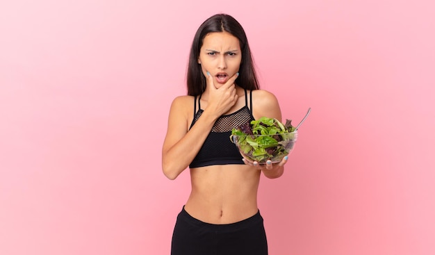 Mujer hispana de fitness con la boca y los ojos bien abiertos y la mano en la barbilla y sosteniendo una ensalada