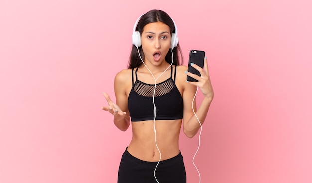 Mujer hispana de fitness que se siente extremadamente conmocionada y sorprendida con auriculares y un teléfono