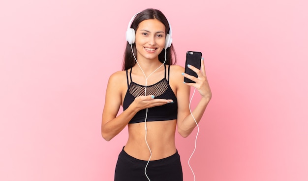 mujer hispana fitness sonriendo alegremente, sintiéndose feliz y mostrando un concepto con auriculares y un teléfono