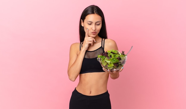 Mujer hispana fitness sonriendo con una expresión feliz y segura con la mano en la barbilla y sosteniendo una ensalada