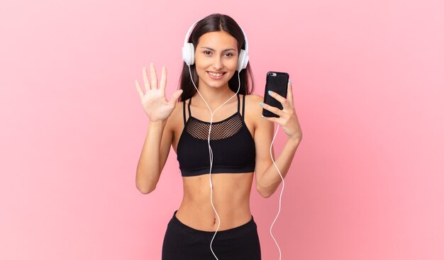 Mujer hispana fitness sonriendo y mirando amigable, mostrando el número cinco con auriculares y un teléfono