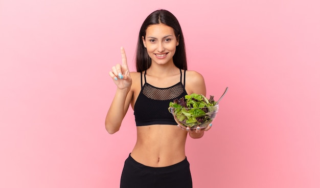 Mujer hispana fitness sonriendo y mirando amigable, mostrando el número uno y sosteniendo una ensalada