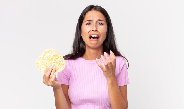 Mujer hispana joven que parece desesperada, frustrada y estresada y que sostiene una galleta de arroz. concepto de dieta