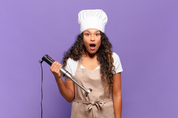 Mujer hispana joven que parece muy consternada o sorprendida, mirando con la boca abierta diciendo wow. concepto de chef