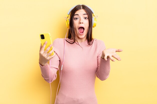 Mujer hispana joven que se siente extremadamente conmocionada y sorprendida. concepto de auriculares y teléfono