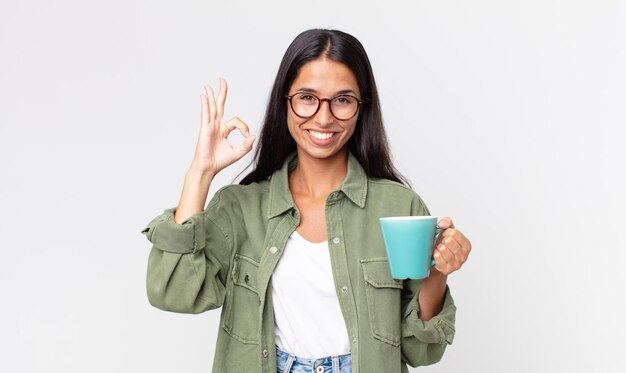 Mujer hispana joven que se siente feliz, mostrando aprobación con un gesto aceptable y sosteniendo una taza de café