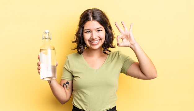 Mujer hispana joven que se siente feliz, mostrando aprobación con gesto bien. concepto de botella de agua