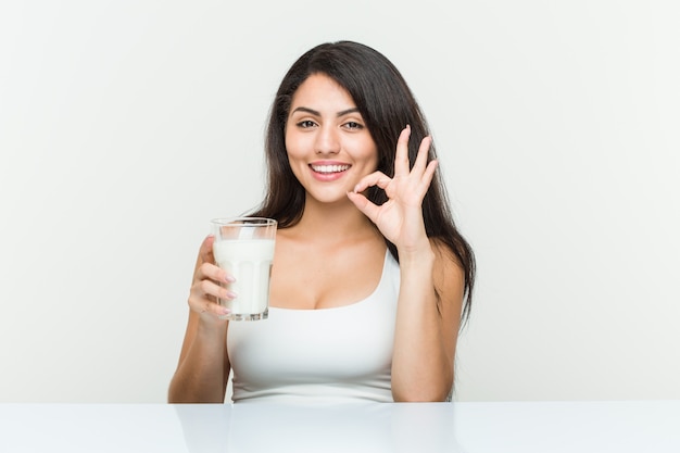 Mujer hispana joven que sostiene un vaso de leche alegre y confidente que muestra gesto aceptable.