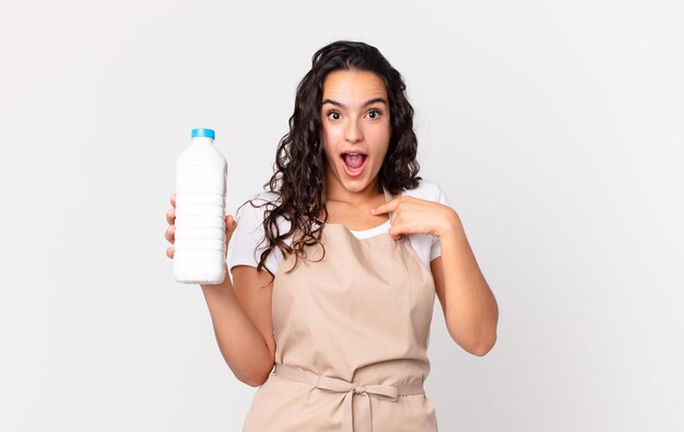 Mujer hispana linda chef que se siente feliz y se señala a sí misma con una emocionada y sosteniendo una botella de leche