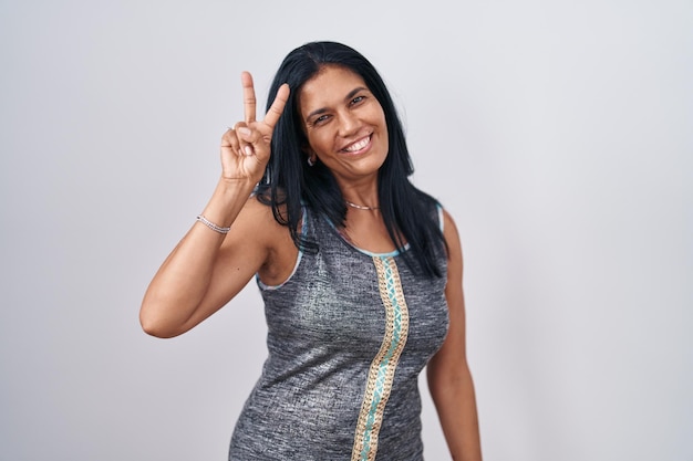 Mujer hispana madura de pie sobre fondo blanco sonriendo mirando a la cámara mostrando los dedos haciendo el signo de la victoria. número dos.