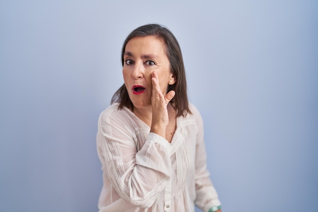 Mujer hispana de mediana edad parada sobre fondo azul con la mano en la boca contando rumores secretos susurrando conversaciones maliciosas