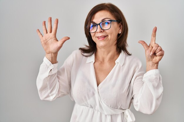 Mujer hispana de mediana edad de pie sobre un fondo aislado mostrando y señalando con los dedos número siete mientras sonríe segura y feliz