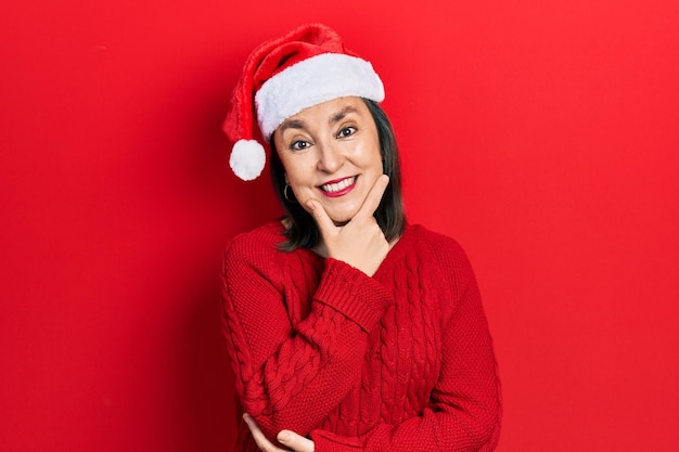 Mujer hispana de mediana edad con sombrero de navidad mirando con confianza a la cámara sonriendo con los brazos cruzados y la mano levantada en la barbilla. pensando en positivo.