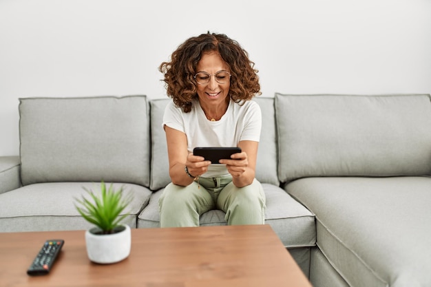 Mujer hispana de mediana edad sonriendo confiada usando un teléfono inteligente en casa