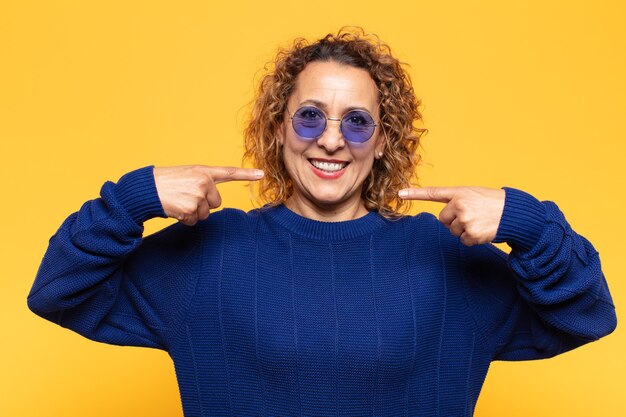 Mujer hispana de mediana edad sonriendo con confianza apuntando a su propia sonrisa amplia, actitud positiva, relajada y satisfecha