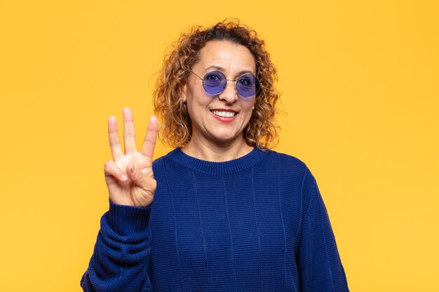 Mujer hispana de mediana edad sonriendo y mirando amigablemente, mostrando el número tres o tercero con la mano hacia adelante, contando hacia atrás