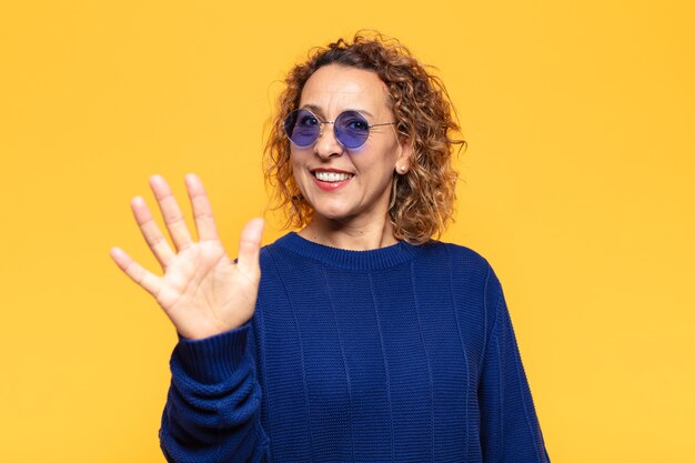 Mujer hispana de mediana edad sonriendo y mirando amistosamente, mostrando el número cinco o quinto con la mano hacia adelante, contando hacia atrás