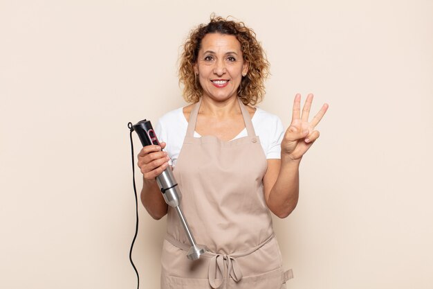 Mujer hispana de mediana edad sonriendo y mirando amistosamente, mostrando el número tres o tercero con la mano hacia adelante, contando hacia atrás