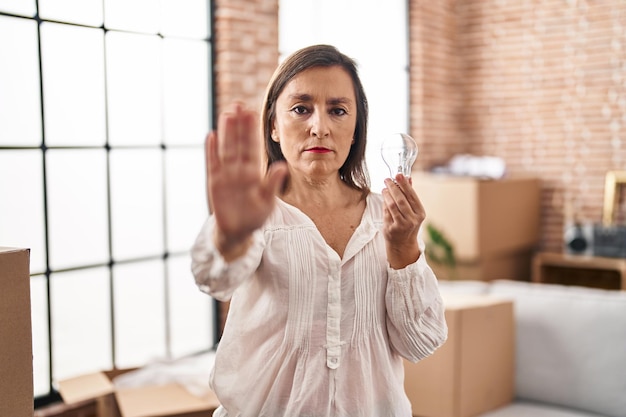 Mujer hispana de mediana edad sosteniendo una bombilla en busca de inspiración e idea con la mano abierta haciendo señas de alto con expresión seria y segura, gesto de defensa