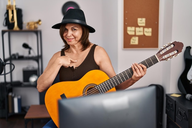 Mujer hispana de mediana edad tocando la guitarra clásica en el estudio de música apuntándose con el dedo a uno mismo sonriendo feliz y orgulloso