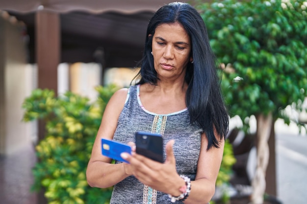 Mujer hispana de mediana edad usando teléfono inteligente y tarjeta de crédito con expresión seria en la calle