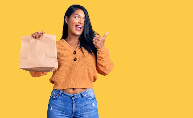 Mujer hispana con el pelo largo sosteniendo una bolsa de papel para llevar apuntando con el pulgar hacia el lado sonriendo feliz con la boca abierta