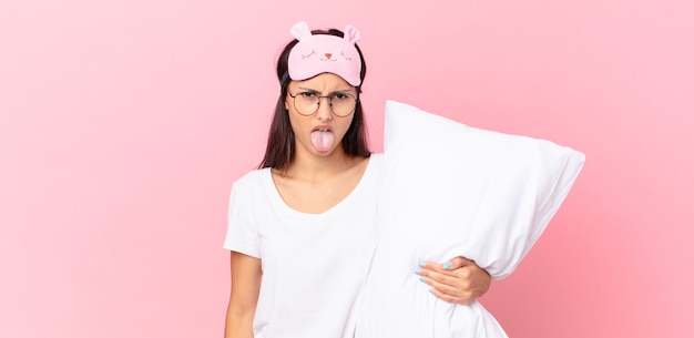 Mujer hispana en pijama sintiéndose disgustada e irritada y con la lengua fuera y sosteniendo una almohada