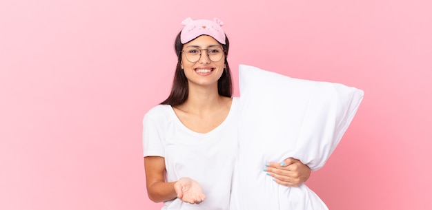 Mujer hispana en pijama sonriendo felizmente con amigable y ofreciendo y mostrando un concepto y sosteniendo una almohada