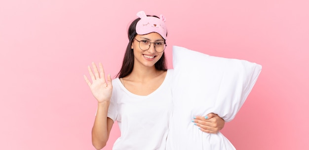Mujer hispana en pijama sonriendo felizmente, saludando con la mano, dándote la bienvenida y saludándote y sosteniendo una almohada