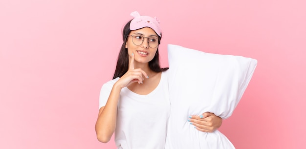 Mujer hispana en pijama sonriendo felizmente y soñando despierto o dudando y sosteniendo una almohada