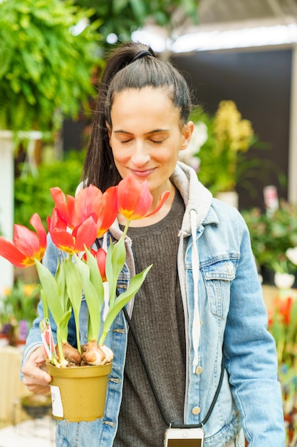 Mujer hispana positiva con cabello negro y ojos cerrados sosteniendo una olla con tulipanes rojos en flor mientras estaba de pie en una tienda de flores ligeras