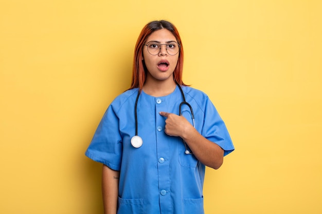 Mujer hispana que parece conmocionada y sorprendida con la boca abierta apuntando al concepto de autoenfermera