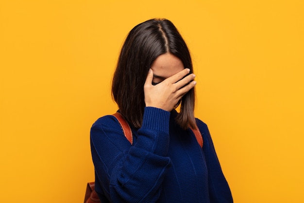 Mujer hispana que parece estresada, avergonzada o molesta, con dolor de cabeza, cubriéndose la cara con la mano