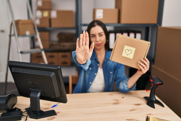Foto mujer hispana que trabaja en una pequeña empresa de comercio electrónico sosteniendo una caja con la mano abierta haciendo una señal de alto con un gesto de defensa de expresión seria y segura